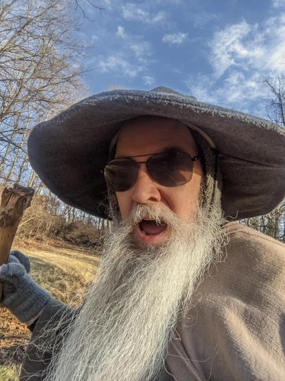 Middle aged man with long grey beard and Gandalf style outfit with hat & staff  wearing sunglasses looks into the camera with a funny/excited look