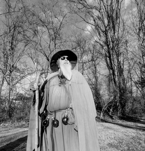 Middle aged man with long grey beard and Gandalf style outfit with hat & staff,  wearing sunglasses, stands in wooded clearing looking off to the right in a black & white photo