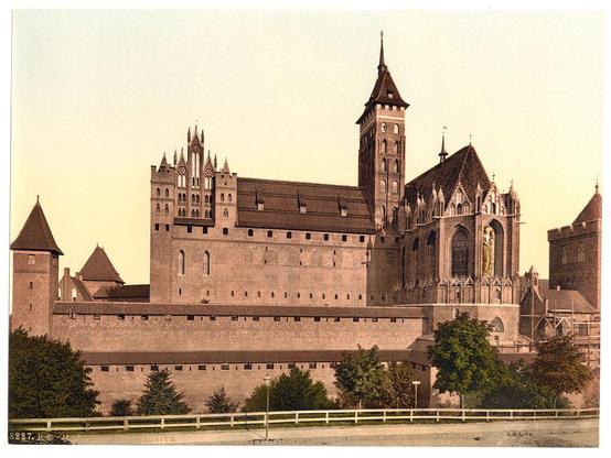 The image depicts an old, historical castle with Gothic architectural features such as pointed arches and flying buttresses. The structure has a fortified appearance with tall walls and towers that suggest defensive purposes in its design. Notable elements include the main building's large arched window adorned with intricate tracery, which likely housed significant religious or royal figures at one time due to its grandeur.

The castle is set against an overcast sky with warm tones of yellow and orange suggesting either sunrise or sunset lighting. The scene appears aged and somewhat faded in coloration, indicative of a print from the late 19th century. In front of the castle are what seem to be simple wooden fences, providing contrast between historical architecture and its surroundings.

There is no visible human activity within this image; it focuses entirely on the architectural beauty and historical significance of the structure itself. The overall atmosphere exudes an air of quiet majesty and historical depth, inviting contemplation about past times and events that took place in such a formidable setting.