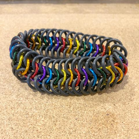 A chainmail bracelet made of European 6-in-1 weave; the outer two rows are black rubber and the inner row is repeating rainbow colours.