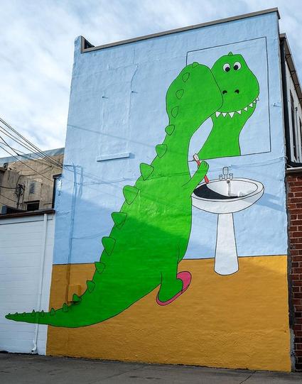 Streetartwall. On the outside wall of a multi-story building is a gigantic mural of a Tyrannosaurus Rex brushing its teeth. It is 35 feet high and 26 feet wide. A cheerfully grinning, cartoon-style green dinosaur stands in front of a bathroom mirror in pink slippers, brushing his teeth. We see him from behind and his face in the mirror. Incidentally, he has lots of square and very pretty teeth.