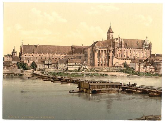 The image depicts a historical scene with Marienburg Castle, also known as Malbork in Poland. The castle is situated by the side of a river that flows beneath several wooden bridges connecting different sections of town across its width. In front of the imposing structure stands another building likely used for administrative purposes or storage during medieval times.

The architecture features classic Gothic elements with large towers and arched windows, typical of the period's design in Prussia (Germany). The overall color tone is sepia-like suggesting that it might be an old photograph from between ca. 1890 to around 1900 as per provided information about its origin.

The river appears calm, with no apparent waves or currents, and a few boats are visible near the bridges indicating some level of activity in transportation across this vital waterway. The sky is clear with scattered clouds suggesting fair weather on that day which could explain why there's such clarity captured by the camera lens used to take this image.

The presence of wooden houses along one side of the river indicates a close-knit community living near their fortified castle, possibly for defense or economic reasons as they might be engaged in trade and other activities facilitated through waterways.