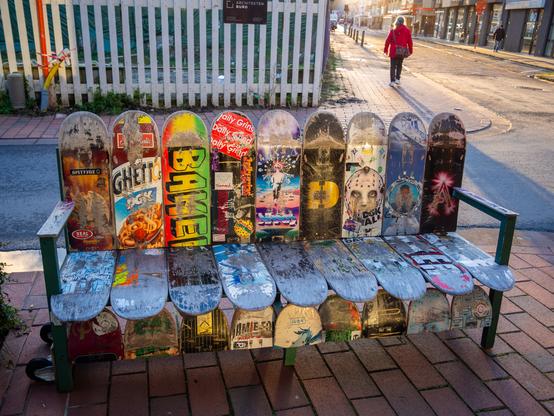 A bench made of skateboards is shown in the image. The bench is made of a metal frame with a wooden top. There are 
various skateboards of different shapes, sizes, and graphics on display. A person is walking away on a paved sidewalk.

Provided by @altbot, generated using Gemini