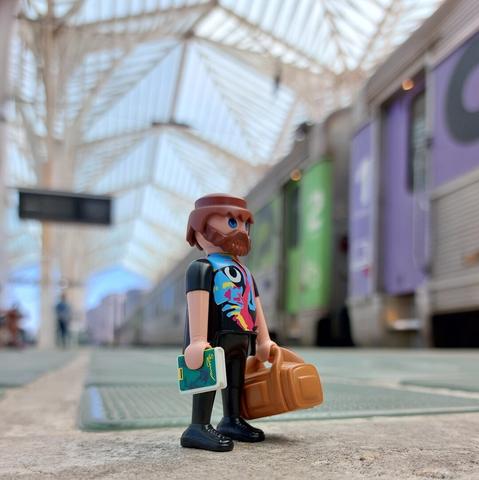 um boneco playmobil parecido com o cantor António Variações, na plataforma da estação Gare do Oriente em Lisboa. No fundo um comboio intercidades com destino a Braga