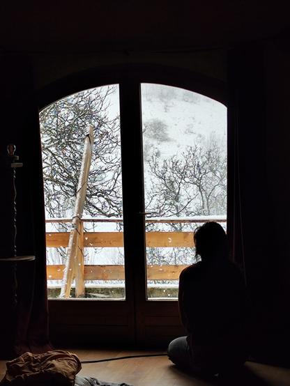 Une personne photographié de dos assise comme pour méditer,  regarde par une fenêtre un paysage enneigé. Le jour et la blancheur de la neige  émanant de la fenêtre contraste avec le clair obscur de la pièce dans un jeu de contre-jour.