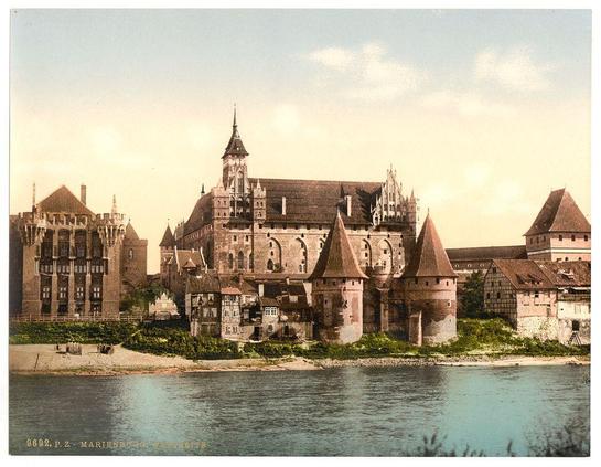 This image depicts a historical castle by the water, likely taken between 1890 and 1900. It's an old photograph of Marienburg Castle in Prussia (now Malbork, Poland). The architecture is characteristic of medieval times with prominent stone towers, turrets, and arched windows. Surrounding buildings appear to be older European-style structures typical for that period. A calm river flows beside the castle grounds, enhancing its picturesque setting.