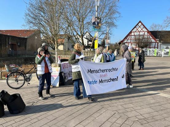 Eine Gruppe Personen steht hinter einem Banner "Menschenrechte statt rechte Menschen". Eine Person redet in ein Mikrofon.