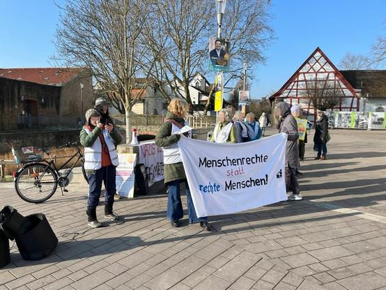 Eine Gruppe Personen steht hinter einem Banner "Menschenrechte statt rechte Menschen". Eine Person redet in ein Mikrofon.