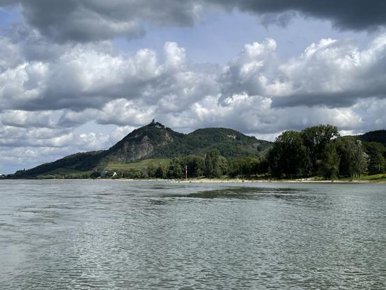 Das Foto zeigt einen Blick über den Rhein auf den Drachenfels, den großen Breiberg und Teile der Insel Grafenwerth (Bad Honnef) im Sommer. Der Himmel ist mit großen Wolken bedeckt und an einigen Stellen scheint blauer Himmel hindurch. Durch die Wolkenlücken bescheint die Sonne die Szenerie teilweise.