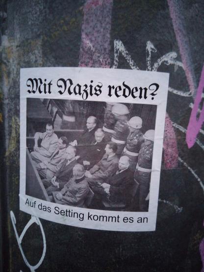 Ein weißer Sticker an einer Ampel: Ein Foto von den Nürnberger Prozessen. Hochrangige Nazis sitzen auf der Anklagebank. Hinter ihnen stehen Gerichtspolizisten der Allierten. Über dem Foto steht in Fraktur: "Mit Nazis reden?". Unten in Antiqua: "Auf das Setting kommt es an".