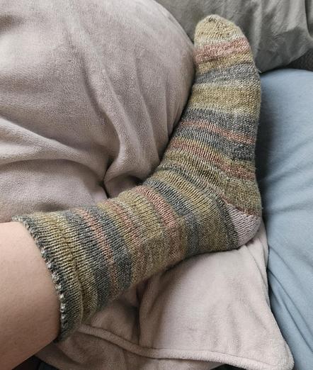 Knit sock on a foot, resting on pale soft pillows. The knit sock is in nature tones of light greens and brown, with a white triangle heel and a scalloped edge.
