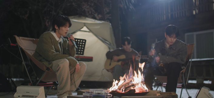 I.N and Seungmin sitting in camp chairs by a fire at night, holding microphones. I.N is singing. Between and behind them, a person plays an acoustic guitar