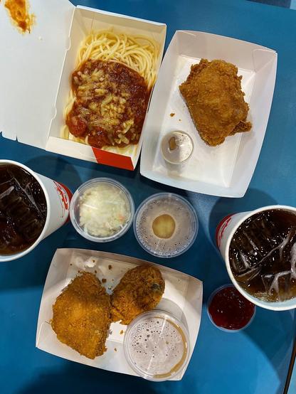 Top down photo of Jollibee food and drinks including a paper plate of 2 Spicy Jollichicken (Fried Chicken) served with a plastic container of gravy, Mashed Potatoes, and Coleslaw, and a combo of a single piece Spicy Jollichicken and a spicy serving of Spaghetti. To the side are 2 cups of Iced Lemon Tea.