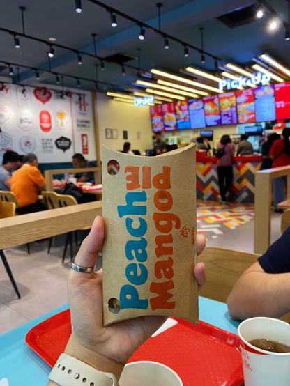 A hand holding a Peach Mango Pie served in a paper container at the forefront, with the Jollibee store and customers seated and eating their food as the background.