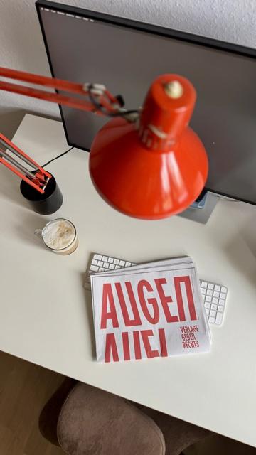 Zeitung auf heller schreibtischplatte. Auf dem Cover steht in rot Augen auf, klein daneben Verlage gegen rechts. Eine knallorangene Lampe und eine Kaffeetasse stehen daneben.