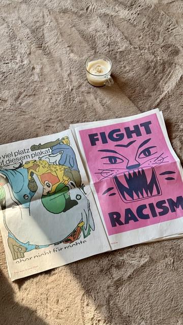Eine aufgeklappte Zeitung liegt auf einem beigen Teppich. Darin zwei bunte Plakate. Auf dem rechten Plakat steht in dunkelblau auf pink fight racism.