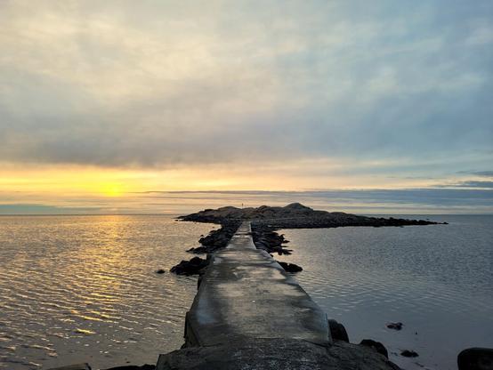 A serene coastal scene at sunset, with a narrow stone pier leading towards rocky islands in the distance. The golden light of the setting sun reflects off the calm water, creating a peaceful atmosphere. The sky transitions from warm hues near the horizon to cooler blues above, with soft clouds stretching across the scene.