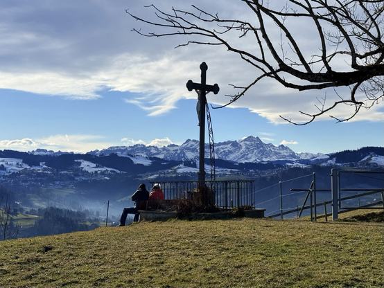 ein kreuz auf dem hügel. darunter geniesst ein paar die aussicht auf den alpstein