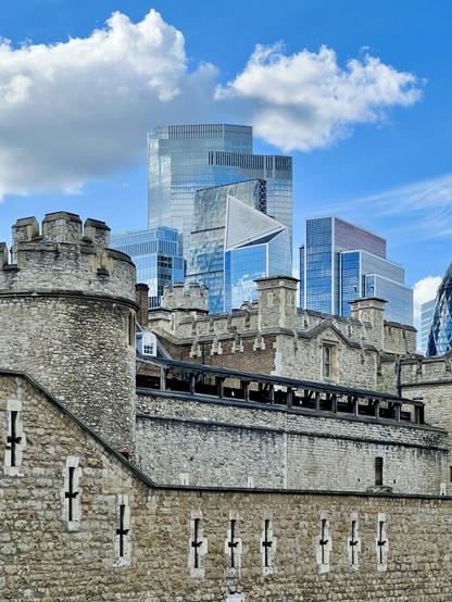 A historic stone castle with battlements in the foreground, contrasted by modern glass skyscrapers in the background under a blue sky with fluffy clouds.