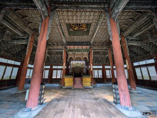 The image depicts the interior of a large, ornate building with a high ceiling and wooden pillars. The walls are adorned with intricate carvings and paintings, while the floor is made of polished wood. In the center of the room, there is a raised platform or stage, which appears to be used for ceremonial purposes. The overall atmosphere suggests that this is a place of great cultural significance and importance.