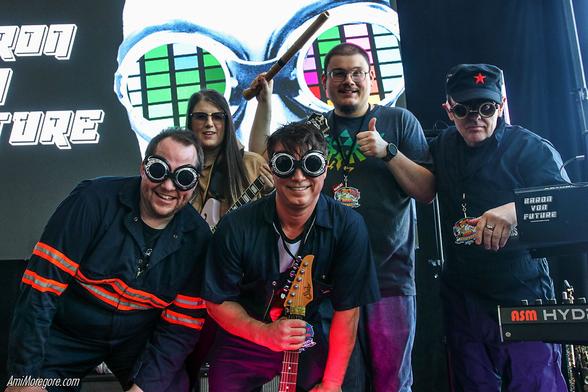 The band Baron Von Future posing together. Three men are wearing goggles. One woman stands behind smiling. One man stands behind holding a flute.