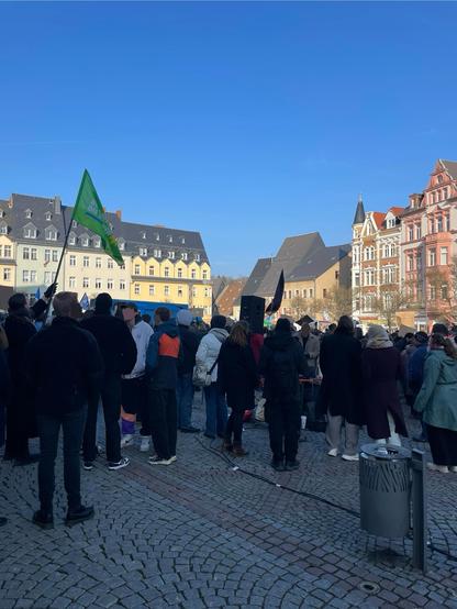 Blauer Himmel, von Sonne beschienene Häuser und viele Menschen. Eine grüne Fahne.