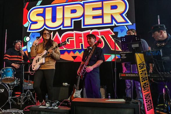 The band assembled on stage with 2 guitarists, a keyboard player and the drummer standing up against a backdrop that says, "Super MAGFest"