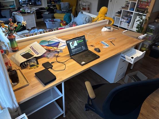 Schreibtisch von schräg oben. Die Platte ist aus Holz. Unter dem Tisch sind ein weißer Schubladenschrank und ein Metallgestell tu sehen.
Auf dem Tisch steht ein Laptop und liegt Zeug.
Vor dem Tisch steht ein Schreibtischstuhl.
Hinter dem Tisch steht ein Sofa aus weißem Holz.
Im Hintergrund ist ein Wohnbereich zu erkennen.