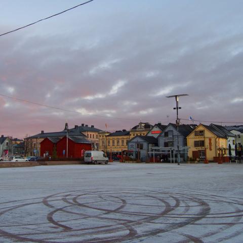A winter scene on the market square in Oulu, Finland. Thick cloud cover in reddish and bluish tones. Light snow cover on the ground has circular car tire tracks on it. The market square is otherwise empty; various buildings can be seen on the far side of the square.