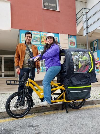 Casal com bicicleta de carga nova amarela e crianças atrás dentro de uma tenda própria para a bicicleta