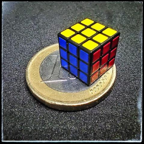 Foto macro de un cubo de Rubik de base negra sobre una moneda de un euro. Se puede ver que ocupa la mitad de la moneda y hasta se aprecia un notable relieve de las pegatinas (no por su grosor fuera de lo normal sino porque el cubo es muy pequeño).
El cubo está resuelto y muestra sus caras amarilla, azul y roja.
