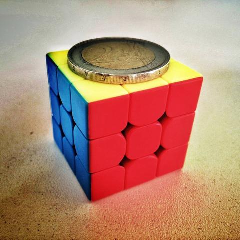 Foto de un cubo stickerless (sin pegatinas, el plástico es del color de cada cara) sobre un fondo claro con textura rugosa.
El cubo está resuelto y podemos ver sus caras amarilla, azul y roja.
Sobre el cubo vemos una moneda de 2 euros que ocupa prácticamente toda la cara amarilla de lado a lado, ya que es casi casi de su mismo tamaño (unos 26 mm).