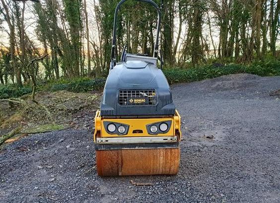 On a gravel-covered clearing with sunlit trees in the background is a small steamroller facing the camera. Its front lights and a cavity between them give it an amusingly frowning face. The roller below this has a lovely rusty brown colour.
