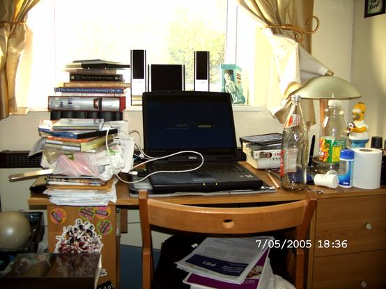 A photograph of a desk. It is very cluttered! In the middle is a black laptop which is open. To the left is a huge pile of books. To the right are several empty bottles.