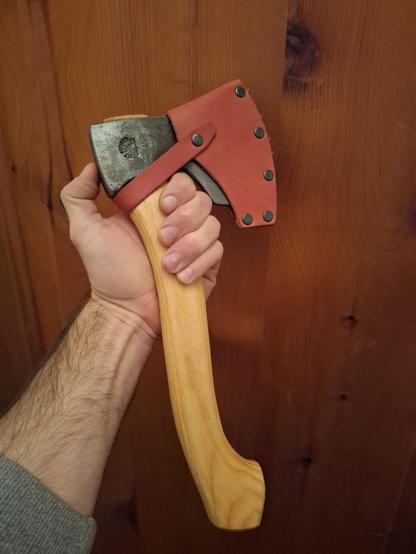 Hand holding small axe. Axe head covered with leather protection. Background oiled arolla pine wood.