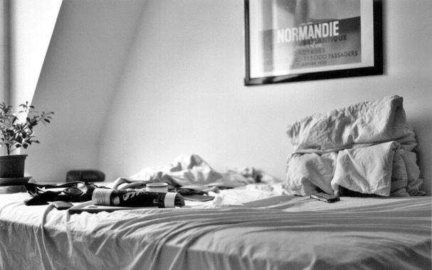Black and white film photography of my bedroom in the early 2000's in Lyon. The bed is undone, there's a coffee cup, an unidentified magazine and, on the wall, a poster of an advert for the cruise boat Le Normandie, by Cassandre in 1935.