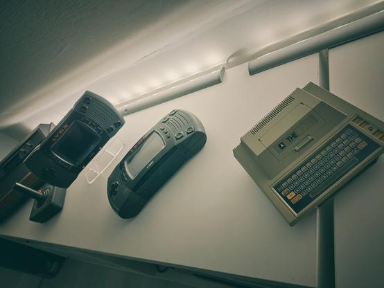 An Atari 400 Mini in the foreground and two Atari Lynx in the background, placed on a shelf