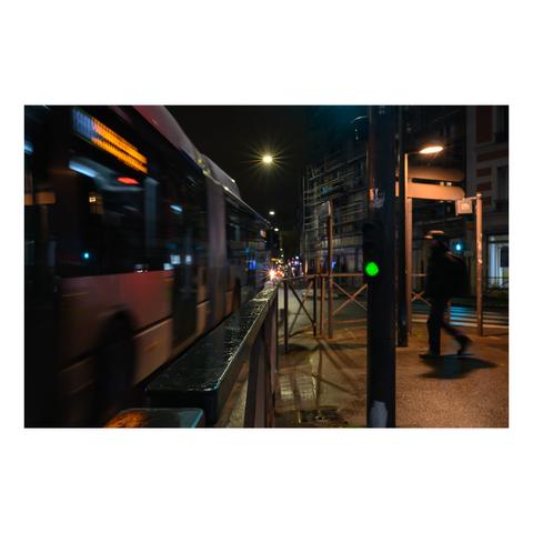 Instantané urbain de nuit avec un bus en mouvement sur la gauche, laissant une traînée de lumière floue. À droite, un passant marche sous un feu piéton vert, dans une rue entourée d'échafaudages et de lampadaires. L'atmosphère est humide, avec des reflets sur les surfaces métalliques et le trottoir.
