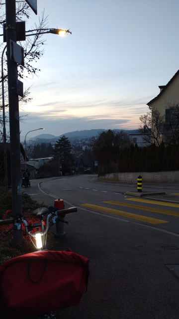 Blick einer an einem Hang bergab Richtung Westen verlaufenden Strasse in die einsetzende Dämmerung. Der Himmel hat einge graue Wolken, aber an vielen Stellen leuchtet der Himmel noch gelb und rosa durch.

Im Vordergrund ein gelber Zebrastreifen und ein an einer Hecke lehnendes Brompton mit roter Fronttasche und grossflächigerer, heller LED-Leuchte am Lenkervorbau. Die Hecke ist vom nicht direkt sichtbaren Rücklicht unterhalb des Sattels rot angeleuchtet.