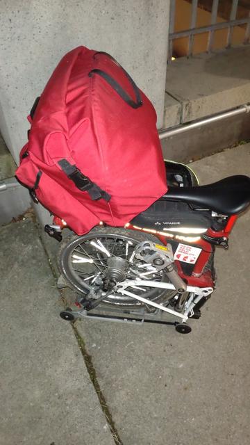 Das gefaltete Brompton steht (nach dem Einkauf) mit vollgepackter, roter Fronttasche vor einem Betonpfeiler.