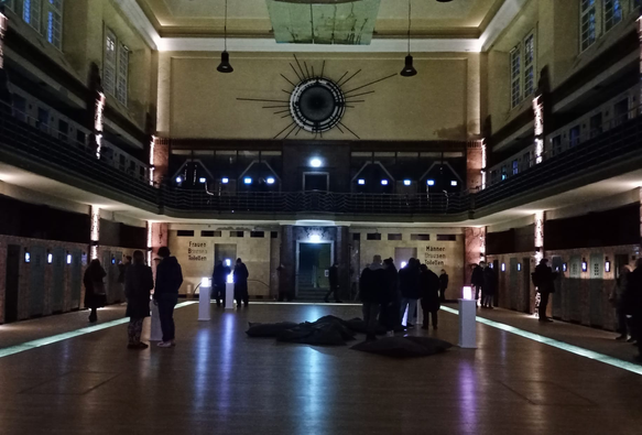 Blick in die alte Schwimmhalle mit Galerie. Das Becken ist abgedeckt und von einem beleuchteten Streifen umgeben. An den Wänden hängen kleine Displays, auf denen die Kunstwerke gezeigt werden. Menschen laufne herum und betrachten die Werke.