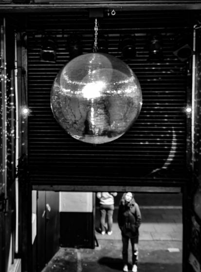 Das Bild zeigt eine große Discokugel, die von der Decke hängt und das zentrale Motiv bildet. Es handelt sich um eine Schwarz-Weiß-Aufnahme, die eine stimmungsvolle, urbane Szene einfangen zu scheint. Im Hintergrund befindet sich ein geschlossenes Rolltor, das eine Art Eingang oder Durchgang andeutet. Darunter steht eine Person, die nach oben zur Discokugel blickt, während eine weitere Person im Hintergrund teilweise sichtbar ist. Der Kontrast zwischen der glänzenden Oberfläche der Discokugel und den dunkleren Elementen der Umgebung verleiht dem Bild eine dramatische und künstlerische Wirkung.