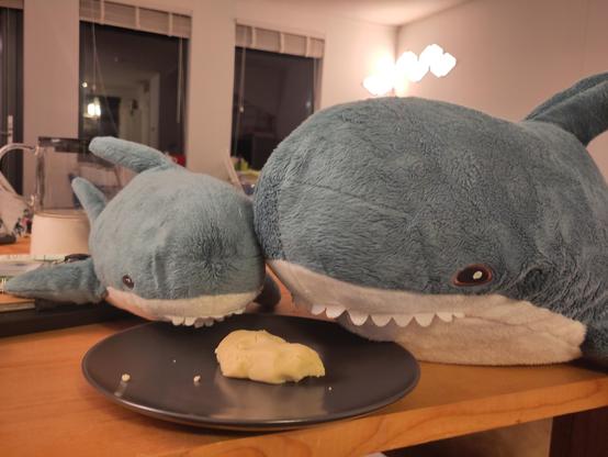 A Smolhaj cuddly shark and a Blåhaj cuddly shark sitting on the table, together behind a plate. On the plate is a leftover of dough.

Don't know what Hapi means with 'the best part', they both got themselves a stomachache!