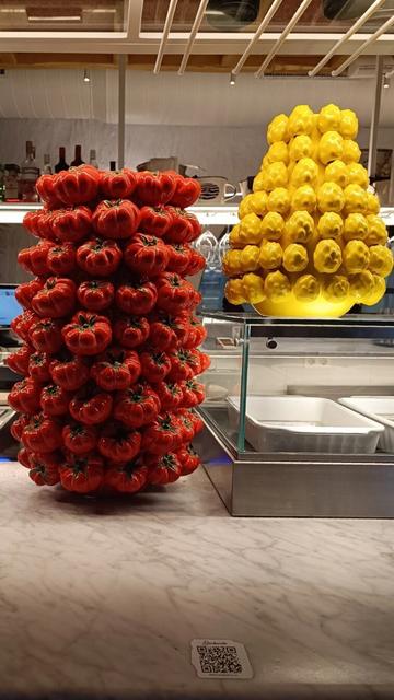 Dos impresionantes jarrones de cerámica italiana expuestos sobre un mostrador de mármol. El jarrón de la izquierda está compuesto por múltiples tomates rojos brillantes con detalles verdes en sus tallos, mientras que el de la derecha presenta una estructura similar, pero con pimientos amarillos.