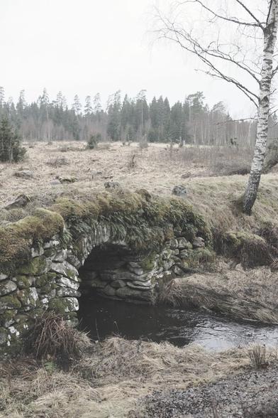 En gammal gräsbevuxen stenbro över ett litet vattendrag. I bakgrunden åkermark och skog mot en grå februarihimmel.
