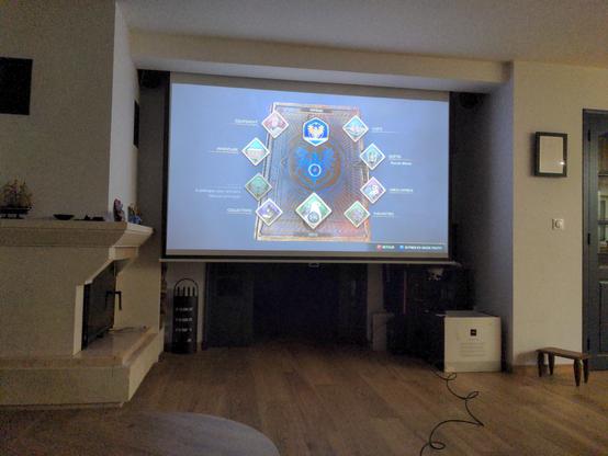 Grand écran de 2,40x1,40m au fond d'une pièce, à côté d'une cheminée sur lequel est projeté le menu du jeu Hogwarts Legacy (Steam via l'image Archlinux HoloISO)