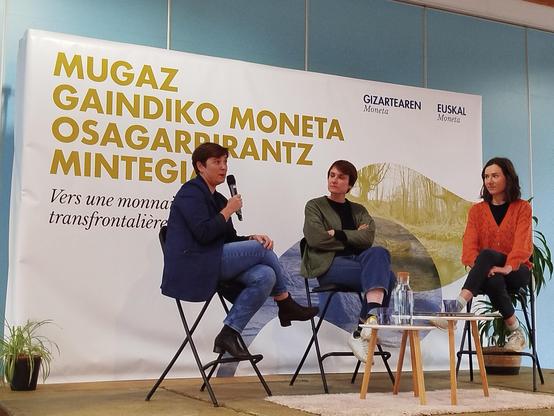 Eszenatokian eserita Aizpea Otaegi hizketan, Cyntia Lamothe eta Lorea Lizarreta, atzean ' Mugaz gaindiko moneta osagarrirantz mintegia' jartzen duen kartel handiarekin.
