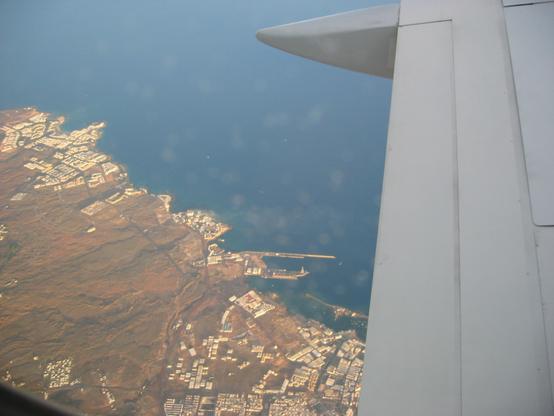 The photo shows an aerial view from an aeroplane over the coastline. The image captures the wing of the plane on the right side, with the sea and a coastal area beneath. The land features a mix of natural terrain and developed areas, including buildings and possibly a harbour or marina along the shoreline.