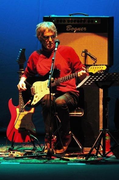 Luis Alberto Spinetta con el cabello canoso y gafas toca una guitarra eléctrica negra sentado en una silla en un escenario iluminado con luz azul. Lleva una camiseta roja y pantalones oscuros. Una guitarra roja está apoyada a su lado. Detrás de él hay un amplificador Bogner. El escenario está iluminado con luces de colores y se ven micrófonos y equipo musical alrededor.