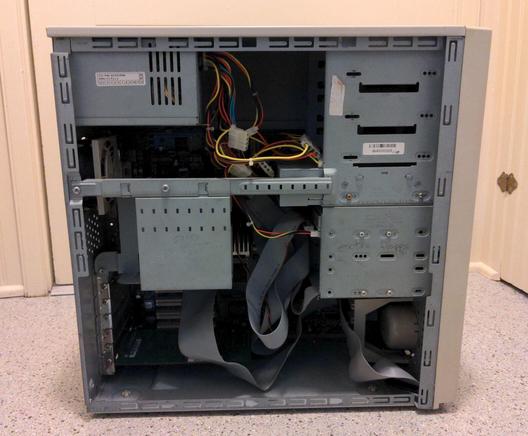 A metal computer case with the side removed to show the PSU at the upper left, front drive bays along the top right, loose power cables, an internal drive bay over the processor, a card in the bottommost PCI slot, and a ribbon cable leading to the top PCI slot, which is hidden by the internal drive bay.